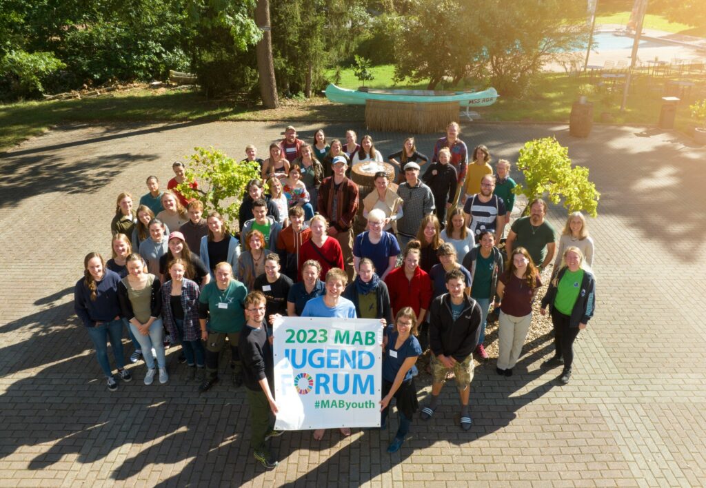 Teilnehmende des MAB Jugendforums gruppieren sich um einen Brunnen und halten ein Banner mit dem Logo des MAB Jugendforums 2023 hoch.