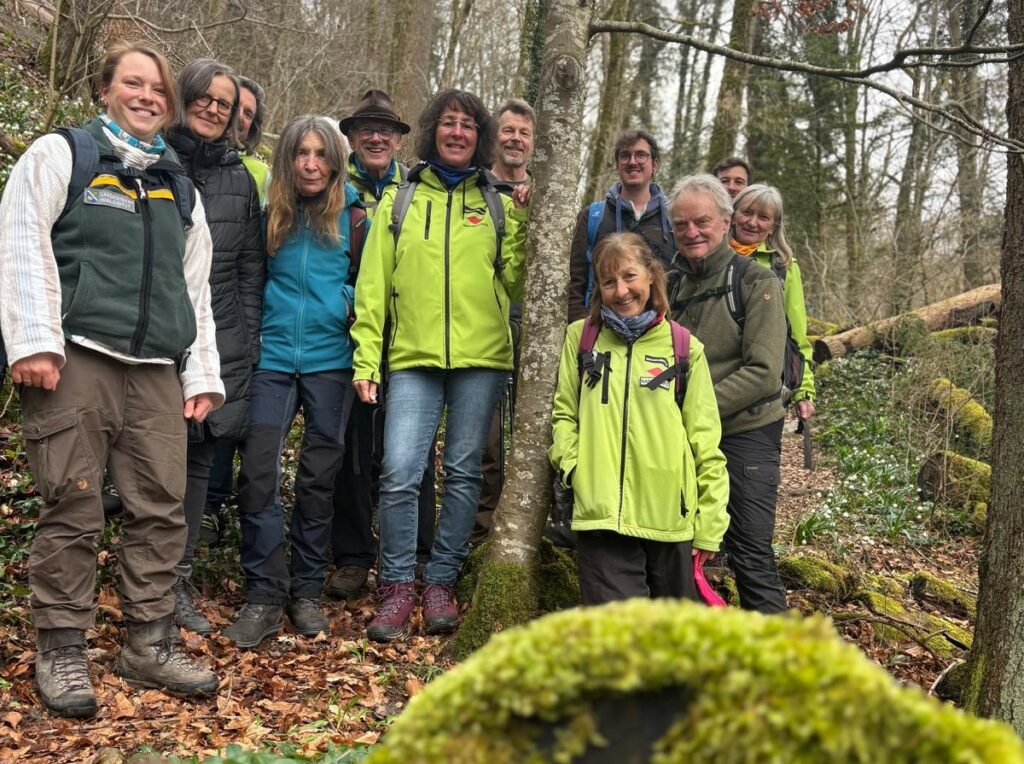 Eine Gruppe Menschen steht eng beieinander im Wald und lacht in die Kamera.