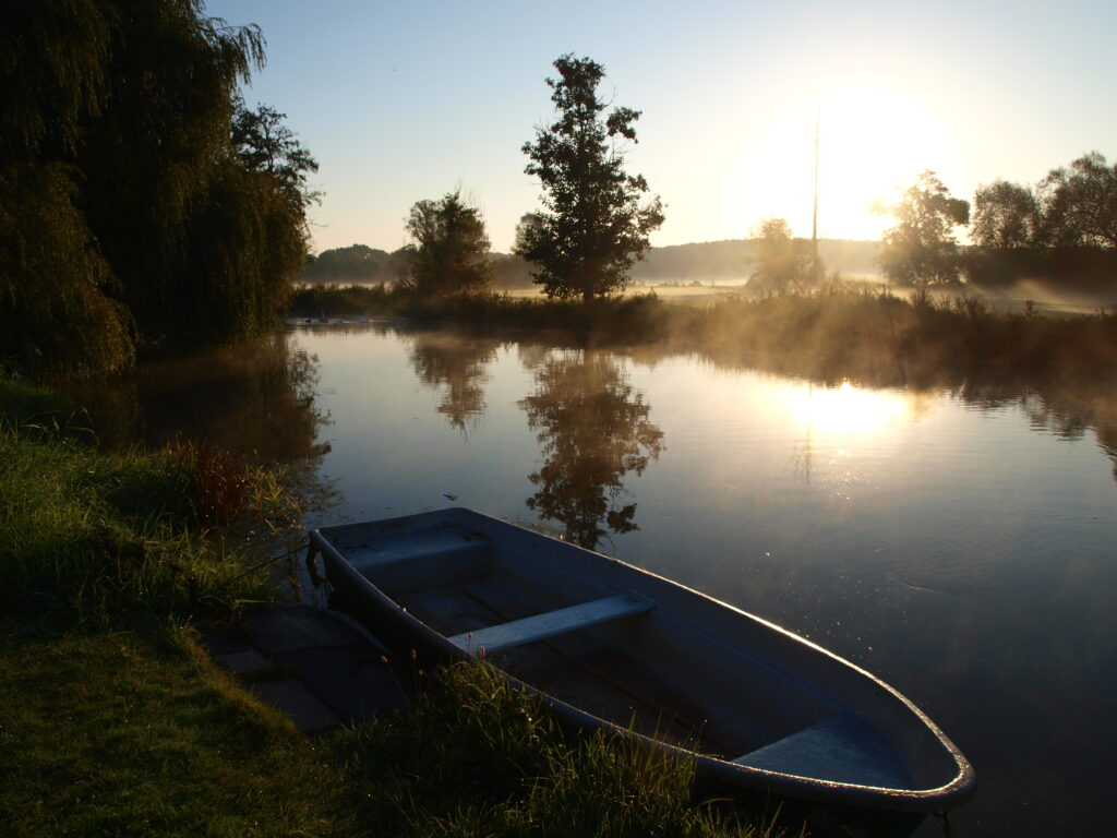 Ein Ruderboot liegt am Fließufer in der Abendsonne. Auf der anderen Uferseite ist Dunst über der Wiese.