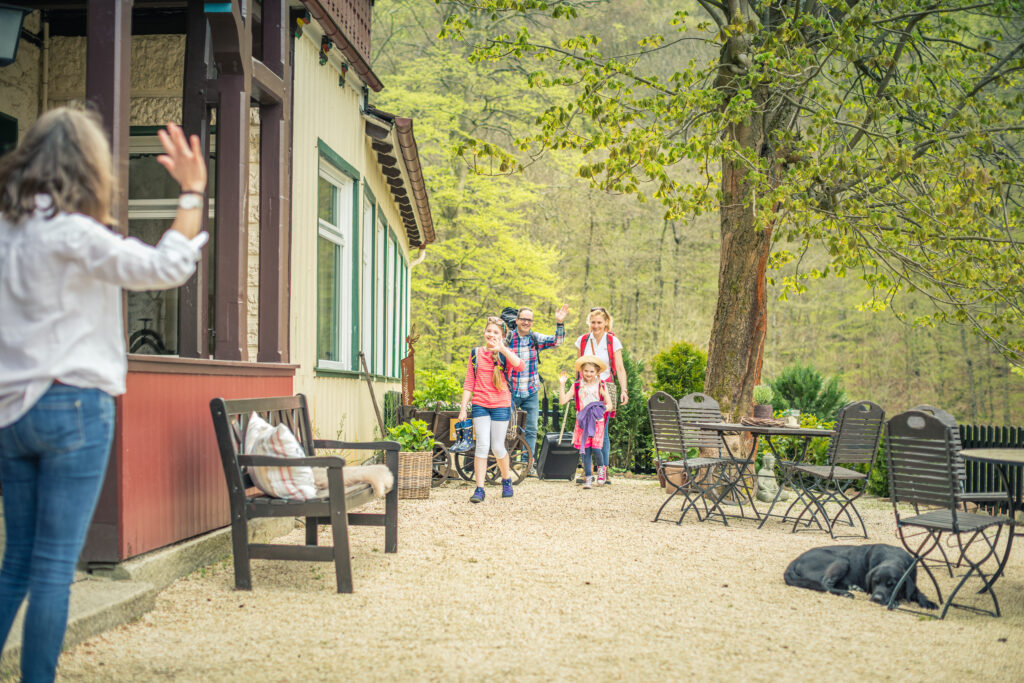Eine Frau winkt einer Familie mit zwei Kindern zu, die mit Gepäck auf dem Hof eines Gasthauses ankommen. Im Vordergrund ruht ein schwarzer Hund entspannt auf dem Boden.