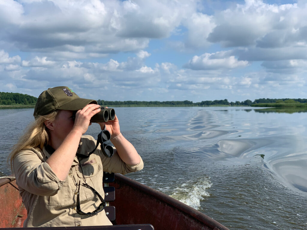 Rangerin sitzt in einem Boot und beobachtet mit dem Fernglas die Umgebung.