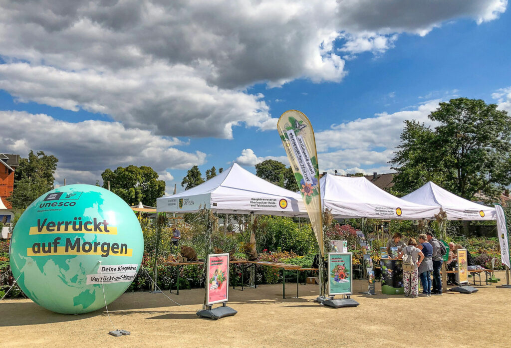 Es sind mehrere Pavillons mit Biosphärenreservatslogo zu sehen sowie ein paar Menschen, die an einem Infostand stehen und ein großer Ball daneben mit der Aufschrift "Verrückt auf Morgen"