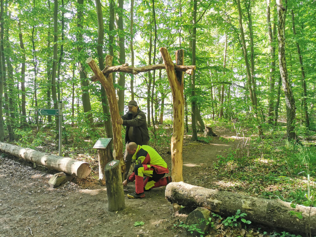 Zwei Personen reparieren einen Torbogen aus Holzstämmen.