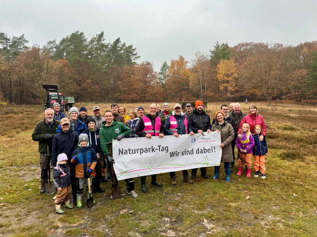 Eine Gruppe Erwachsene und Kinder verschiedenen Alters stehen für ein Gruppenfoto in der Heide und halten ein Banner auf dem "Naturpark-Tag. Wir sind dabei!" steht.