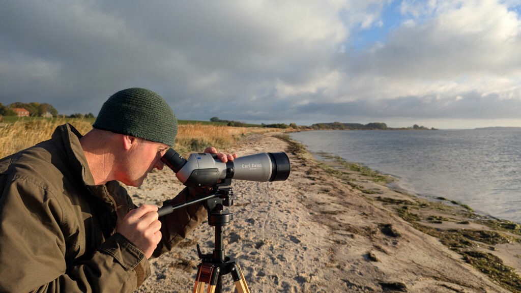 Eine Person steht mit einem Fernglas, stabilisiert durch ein Stativ auf dem Strand und blickt aufs Meer.