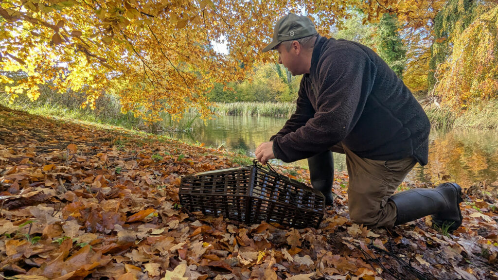 Eine Person hockt auf dem Waldboden, in den Händen ein Reuse. Im Hintergrund ist ein Gewässer erkennbar.