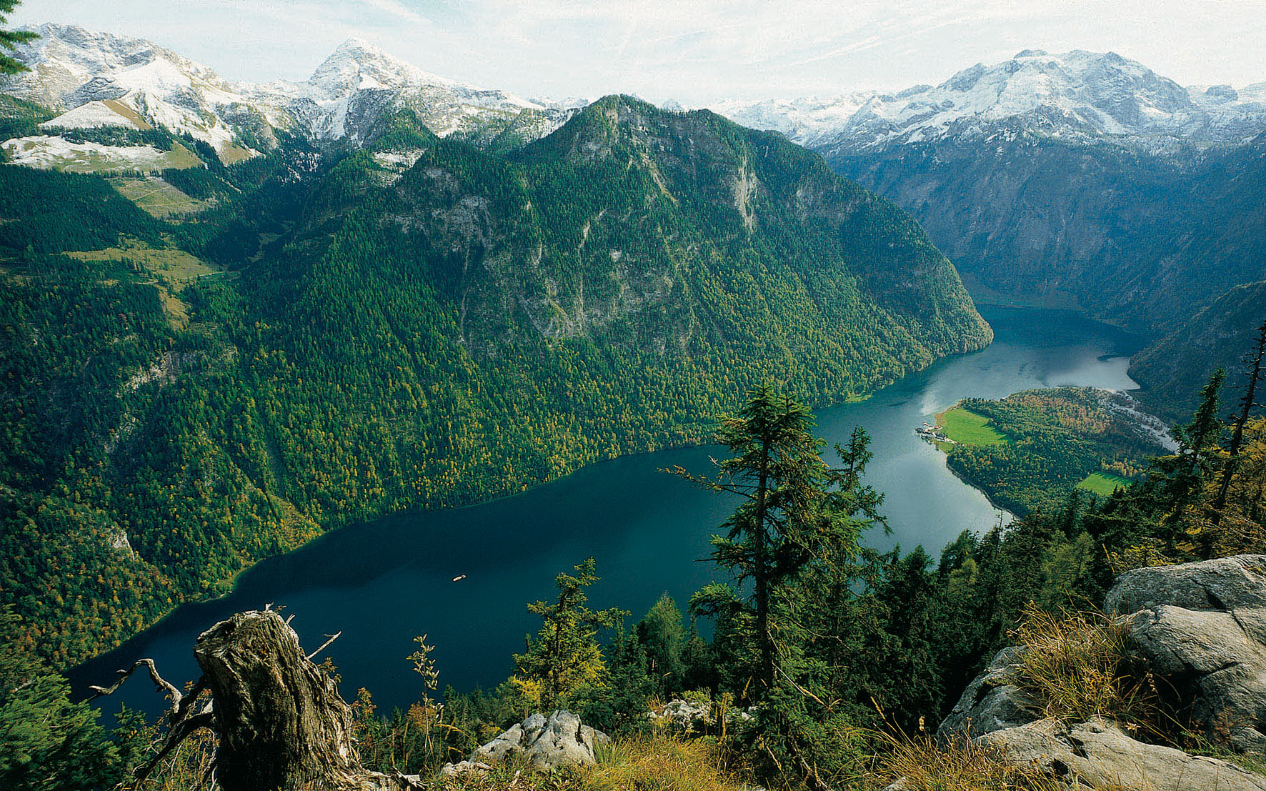 Aussicht von der Archenkanzel herunter auf einen großen See. Der See ist umgeben von steilen und zum Teil bewaldeten Berghängen. Im Hintergrund sind die Spitzen der Berge schneebedeckt.