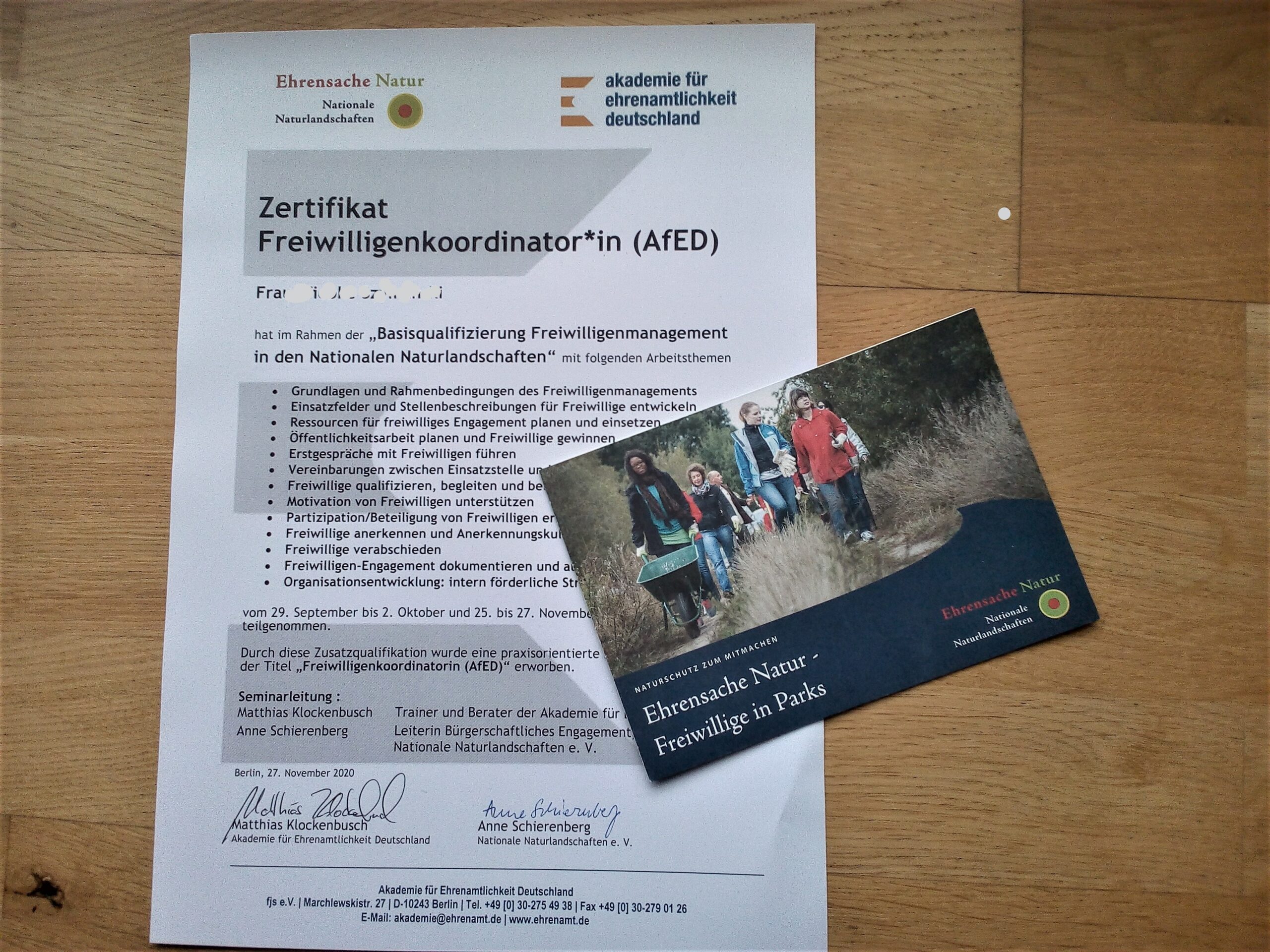Ein Zertifikat für Freiwilligenkoordinator*innen und eine Postkarte, mit Überschrift "Ehrensache Natur - Freiwillige in Parks" und einem Ehrensache Natur Logo in der Ecke.