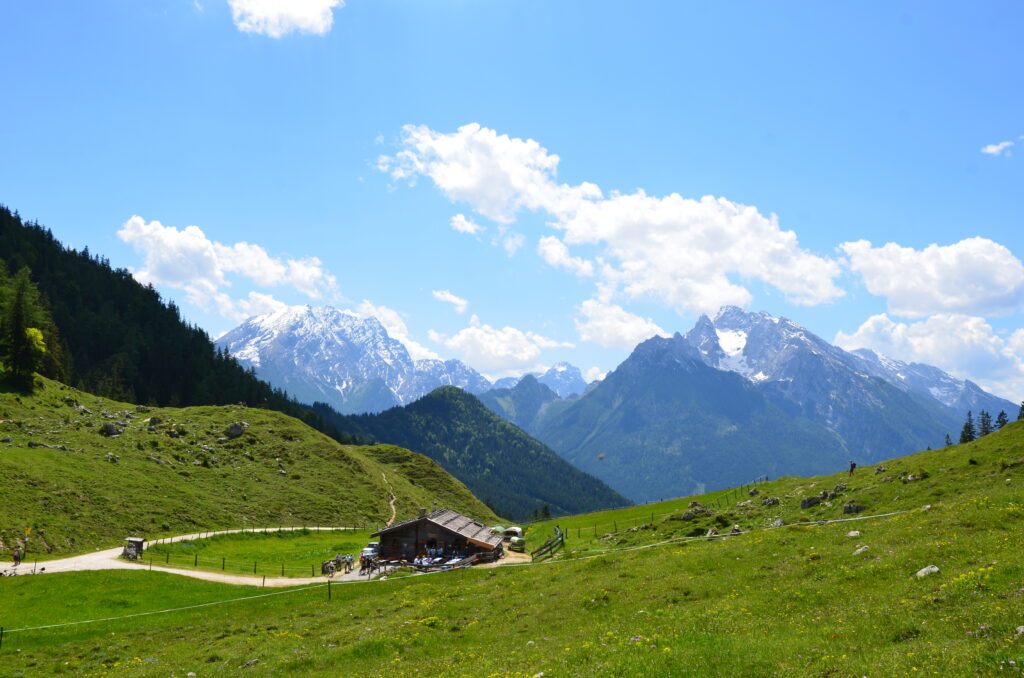 Eine Almhütte mit Blick auf mehrerer Berggipfel. Die Sonne scheint und Wanderer machen auf der Terasse Rast.