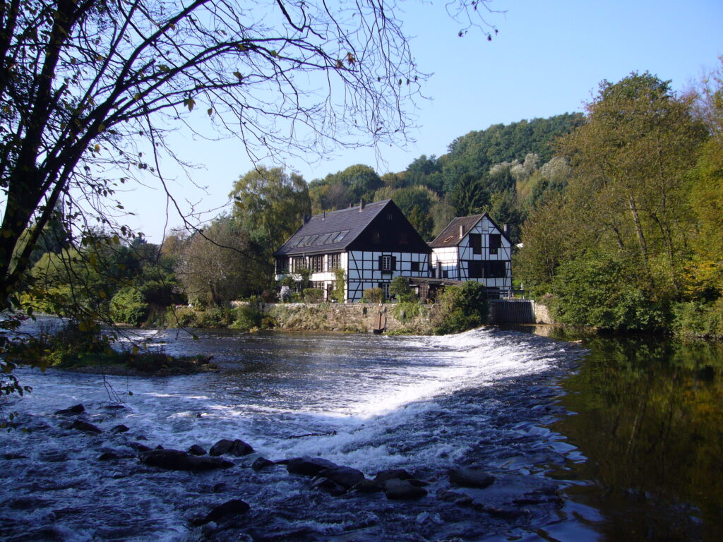 Ein uriges Fachwerkhaus steht an einem sprudelnden Fluss. Um das Haus herum ist grüner Wald.