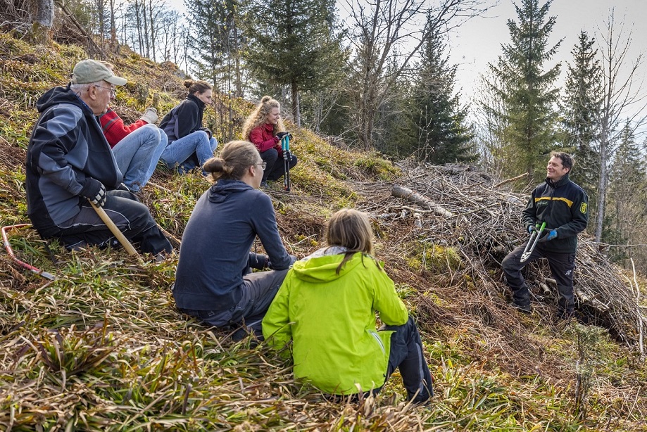 Eine Gruppe mit 6 Erwachsenen sitzt mit Astscheren in der Hand auf einem steilen Hang, vor Ihnen ein Mann stehend mit Astschere, der zur Gruppe spricht. Im Hintergrund Nadelbäume und ein großer Reisighaufen