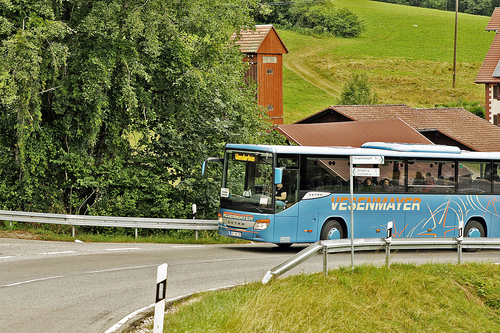 Ein blauer Bus mit der Aufrischrift "Wanderbus" biegt um eine Straßenkurve. Im Hintergrund ist eine grüne Wiese sichtbar.