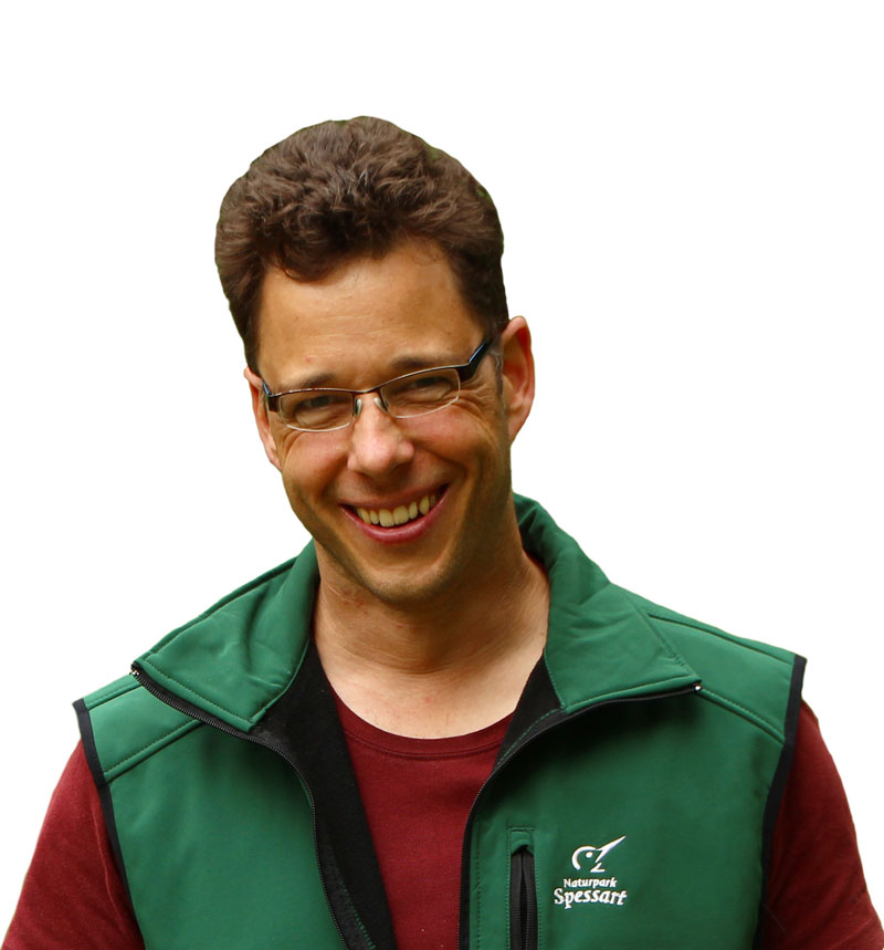 ein Mann mit kurzen braunem Haar, Brille und grüner Weste lächelt in die Kamera